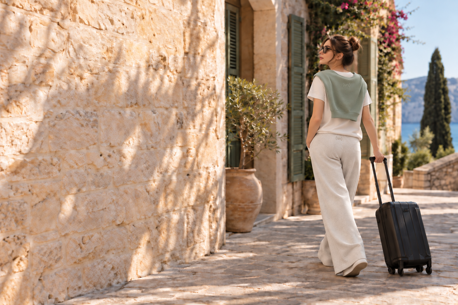 Light-layering concept: a woman in a bone tee and flowing oat pants with a sage crew draped over her shoulders at a warm arrival