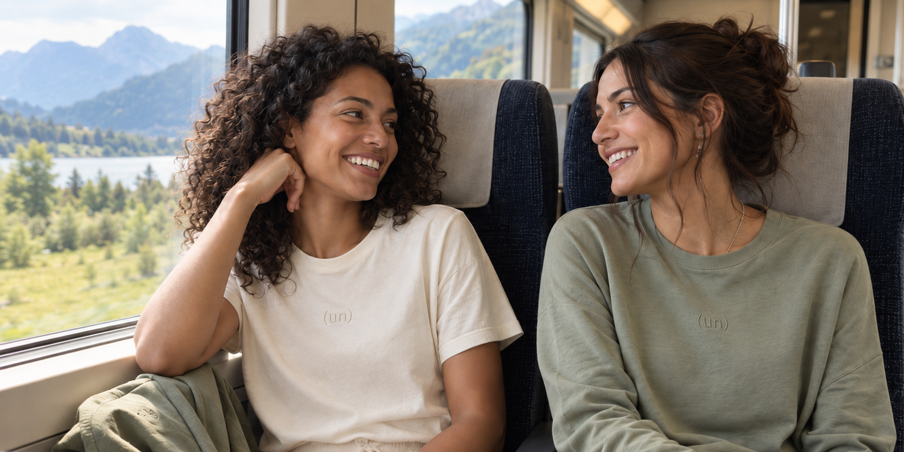 Waist-up travel apparel concept: two adult friends on a train wearing bone and sage tops with discreet (un) marks
